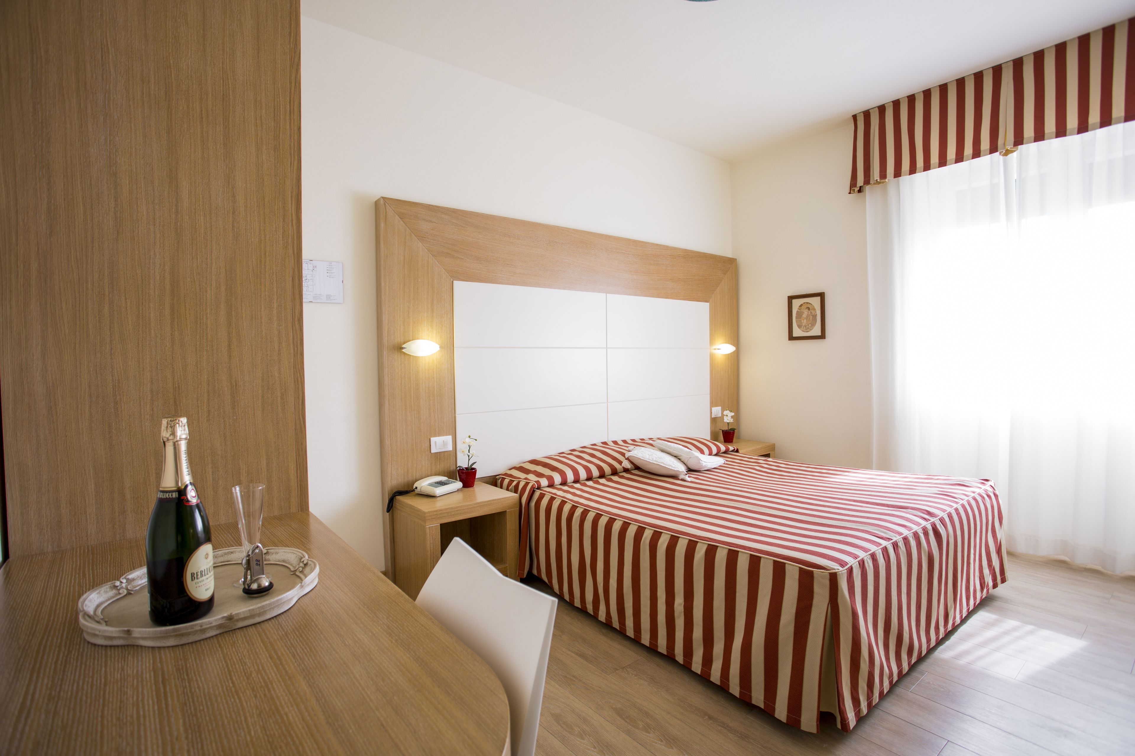 A hotel room with a bed, a bedside table with a bottle and glasses, a chair, and a wooden headboard.