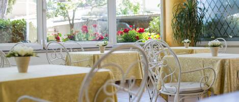 Un espace repas avec des tables recouvertes de nappes jaunes et des chaises en métal blanc, offrant une vue sur un jardin grâce à de grandes fenêtres.