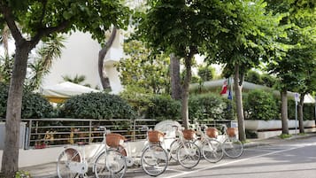 A row of white bicycles with baskets parked along a sidewalk, surrounded by trees and a building with balconies.