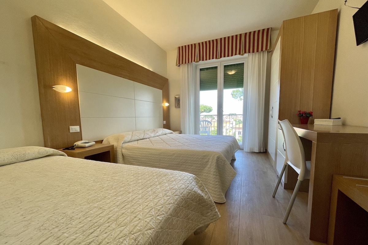 A hotel room with two beds, a desk, a chair, and a balcony with a view.