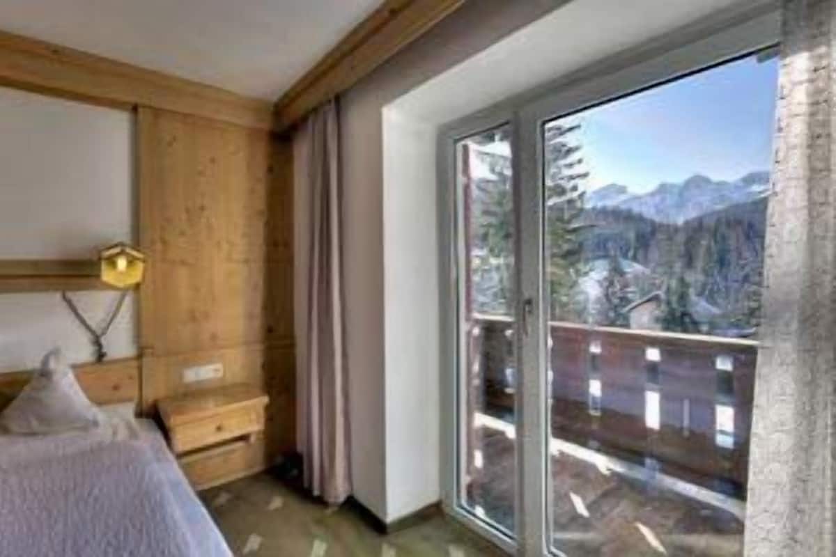 A bedroom with a wooden headboard, a bed with a white blanket, a nightstand, a lamp, and a large window with a mountain view.