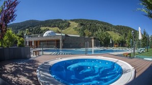 A hot tub on a wooden deck with a pool in the background, a building with a dome, and a mountainous landscape. - Hotel Nordic (El Tarter)