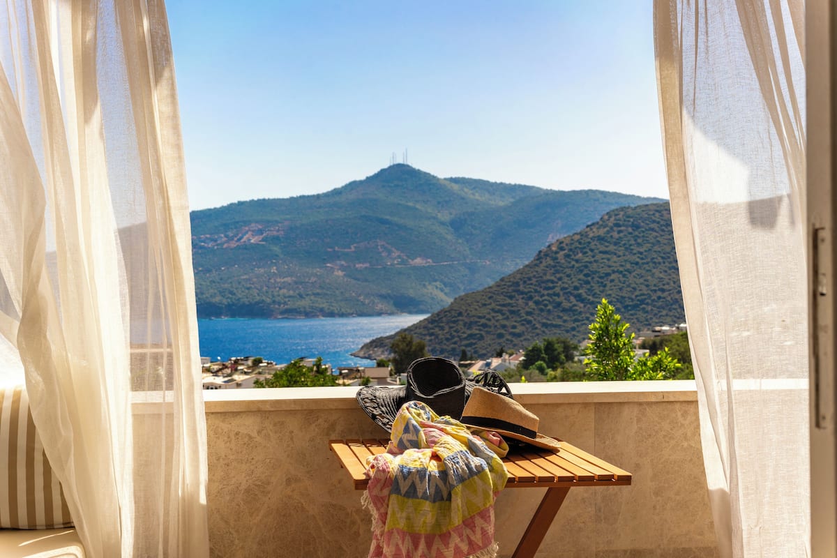 A balcony with a view of the sea and hills, a wooden table with a hat and a bag, and sheer curtains.