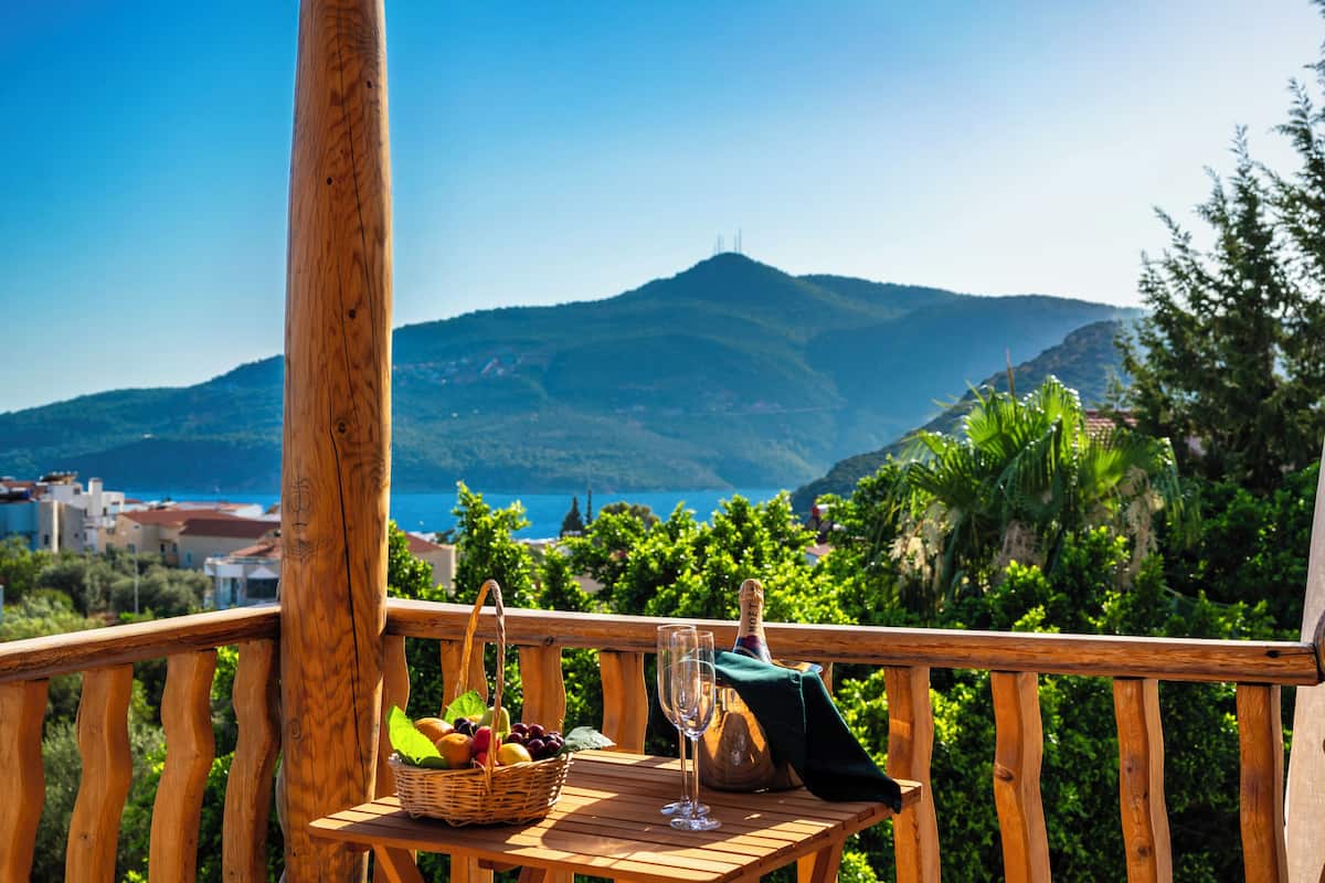 A wooden deck with a table holding a basket of fruits, a wine glass, and a bottle of champagne. The deck overlooks a scenic view of mountains and a body of water.