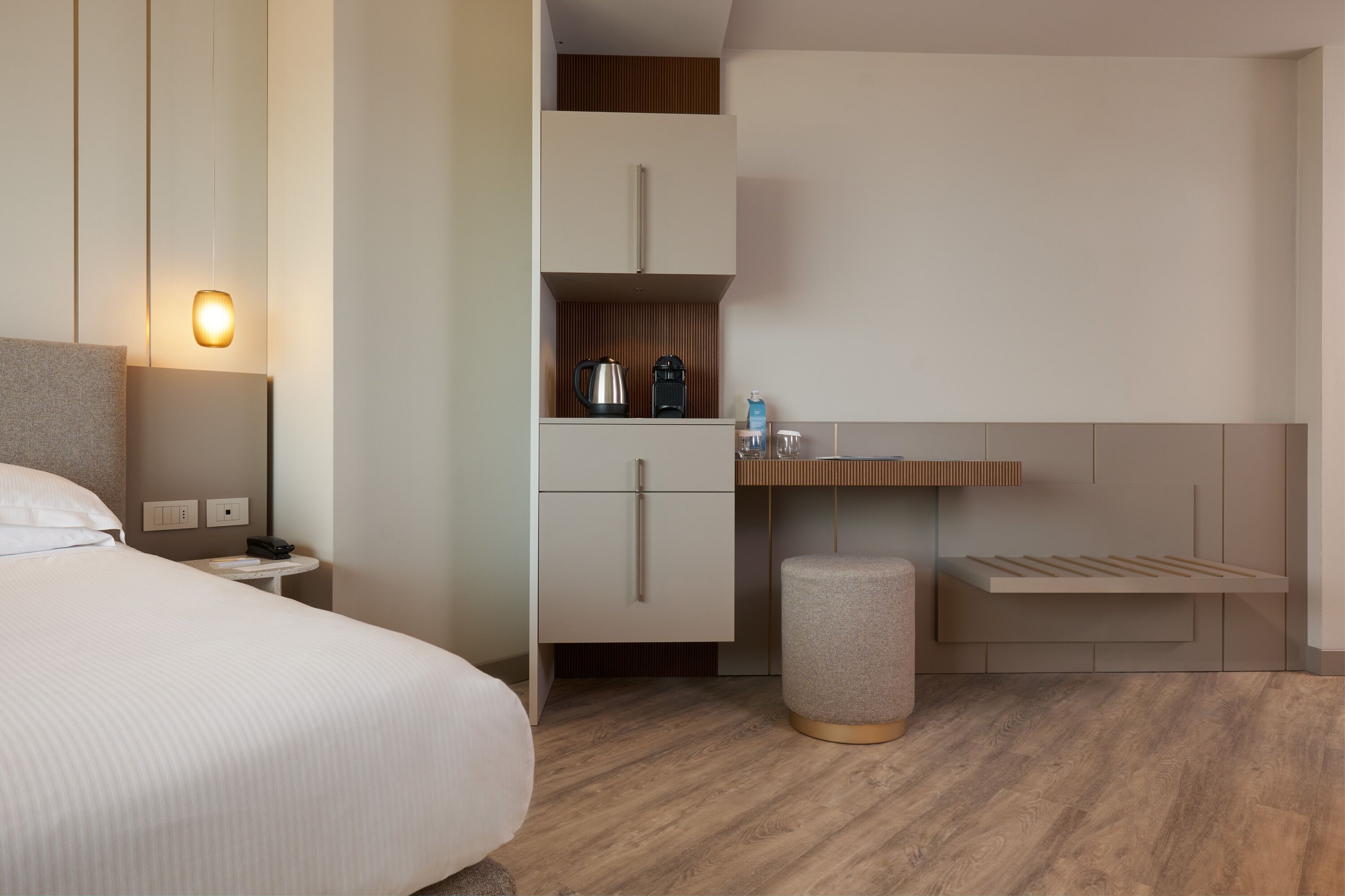A modern hotel room with a bed, bedside table, lamp, built-in wardrobe, and a small stool.