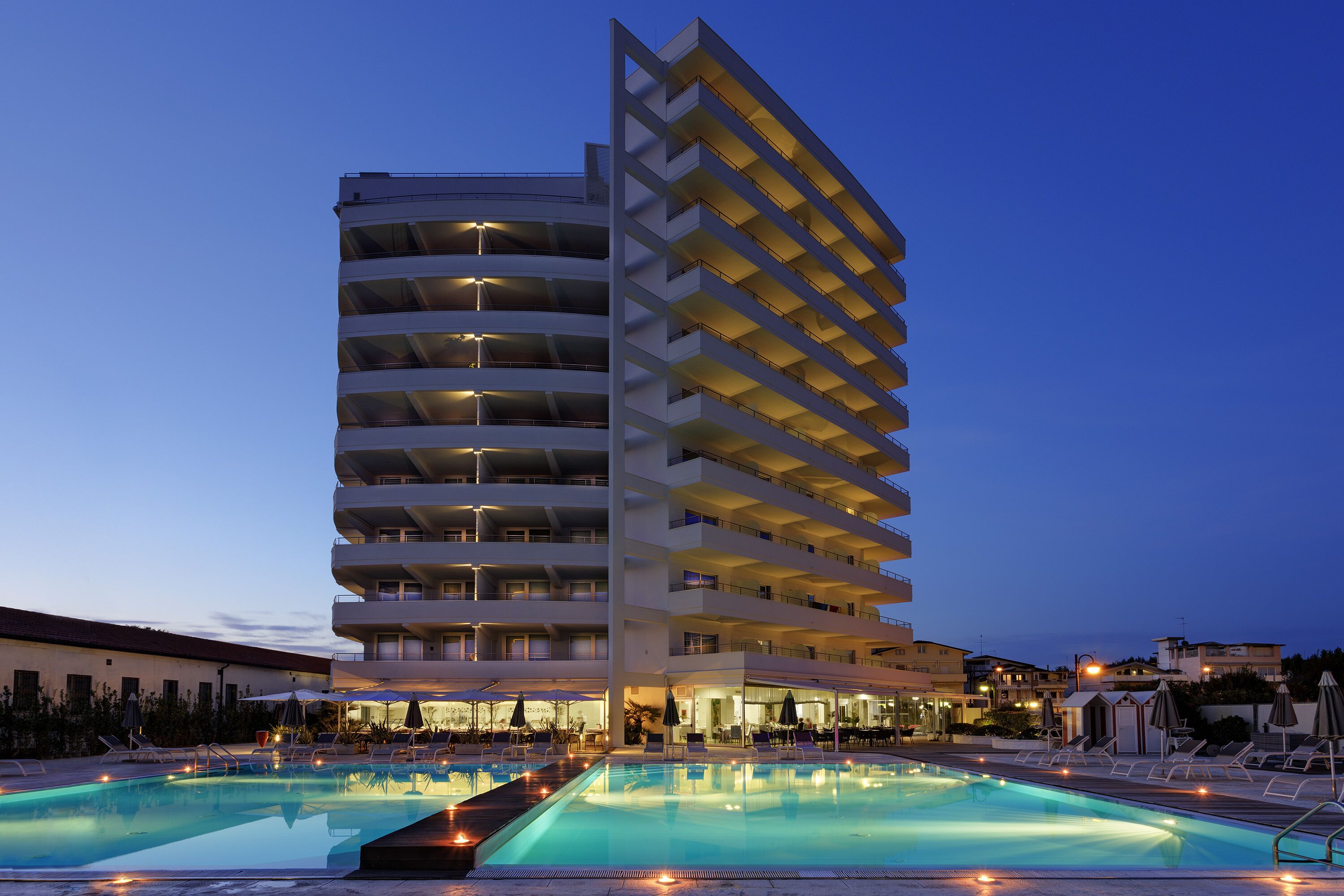 A multi-story hotel with a swimming pool, outdoor seating, and a well-lit facade.