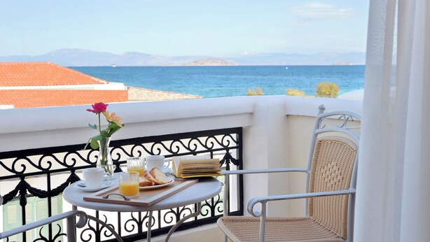 A balcony with a table set for breakfast, a wicker chair, and a view of the sea and mountains. - Hotel Agistri (Agistri)