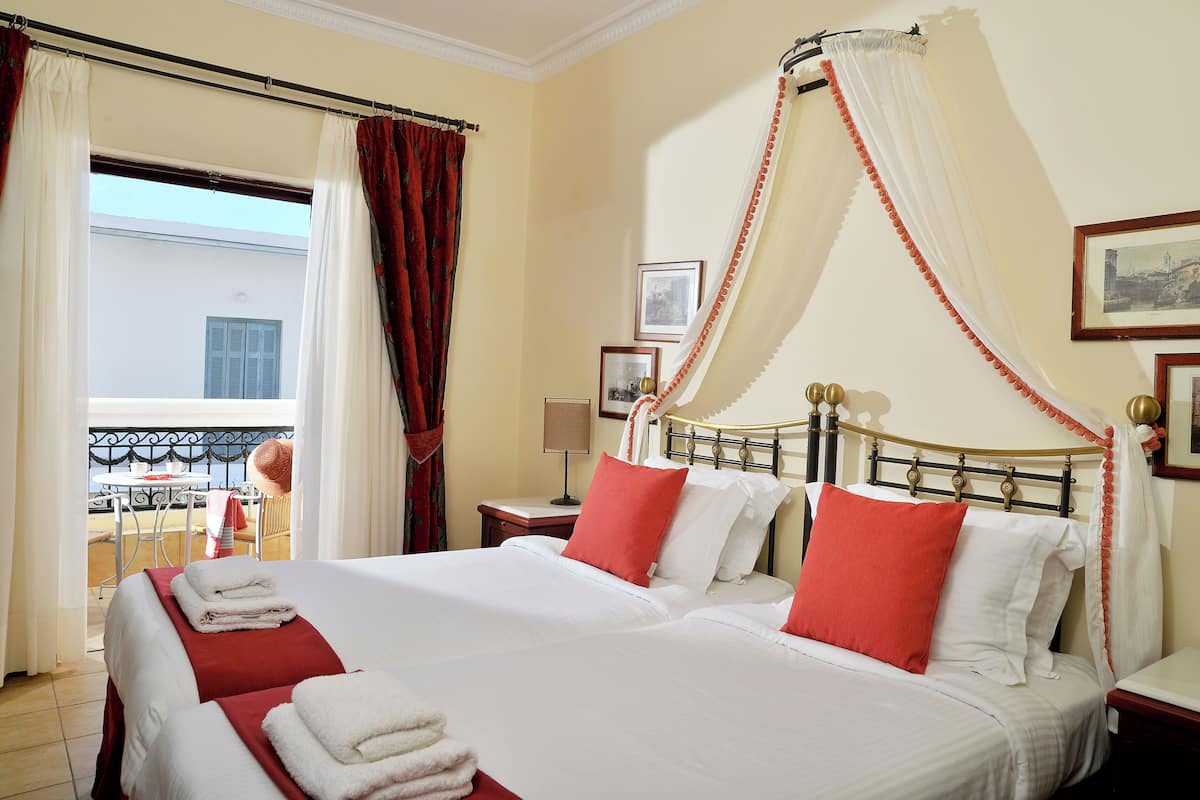 Cama doble con ropa de cama blanca y roja, una mesita de noche con lámpara y un balcón con mesa y sillas.