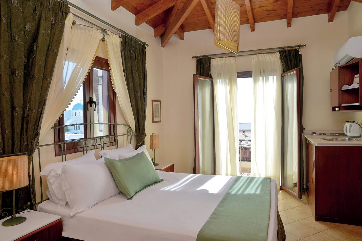 Habitación de hotel con cama, mesita de noche, lámpara, ventana con cortinas, puerta a un balcón y una pequeña cocina.