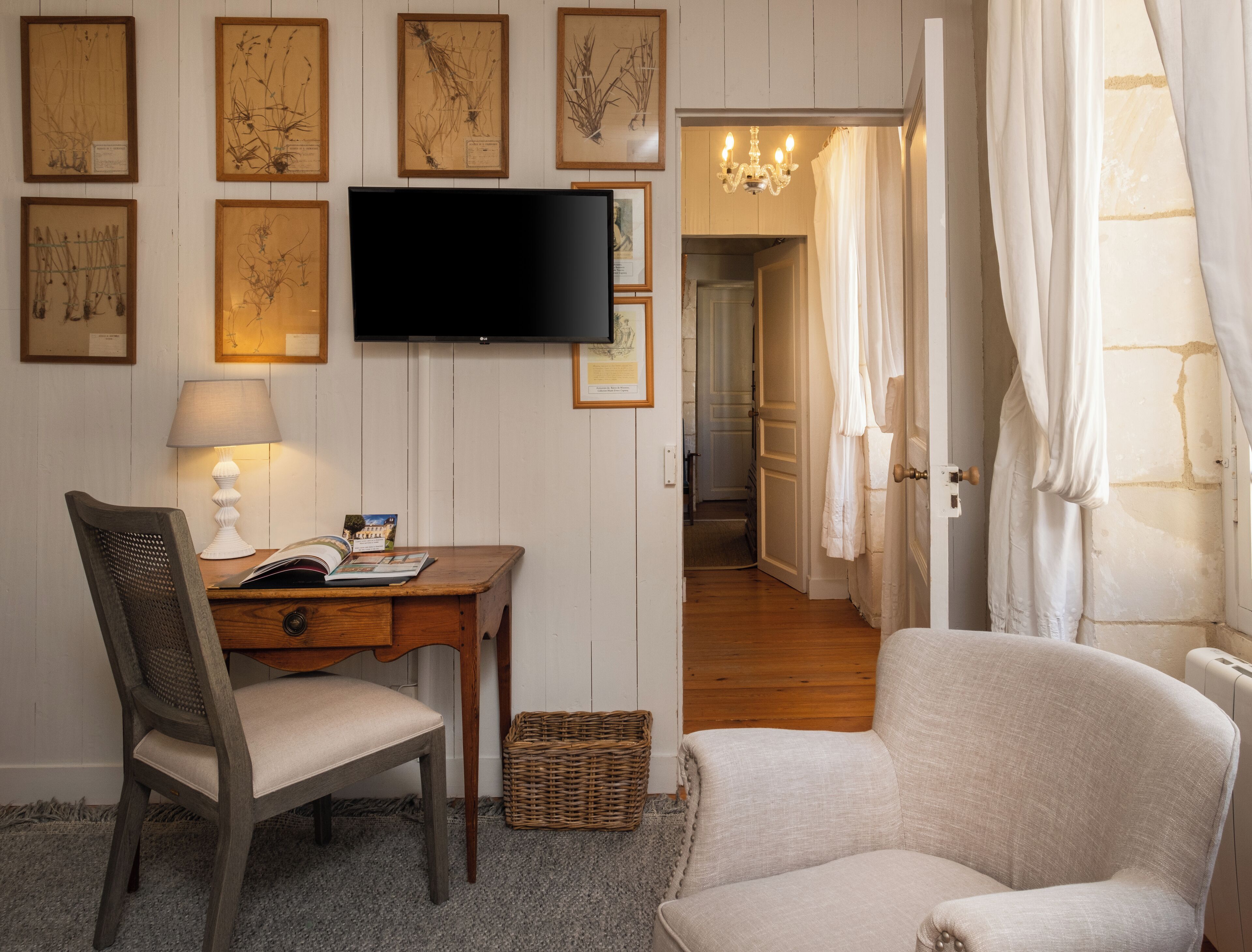 A room with a wooden desk, a chair, a television, and framed botanical prints on the wall.