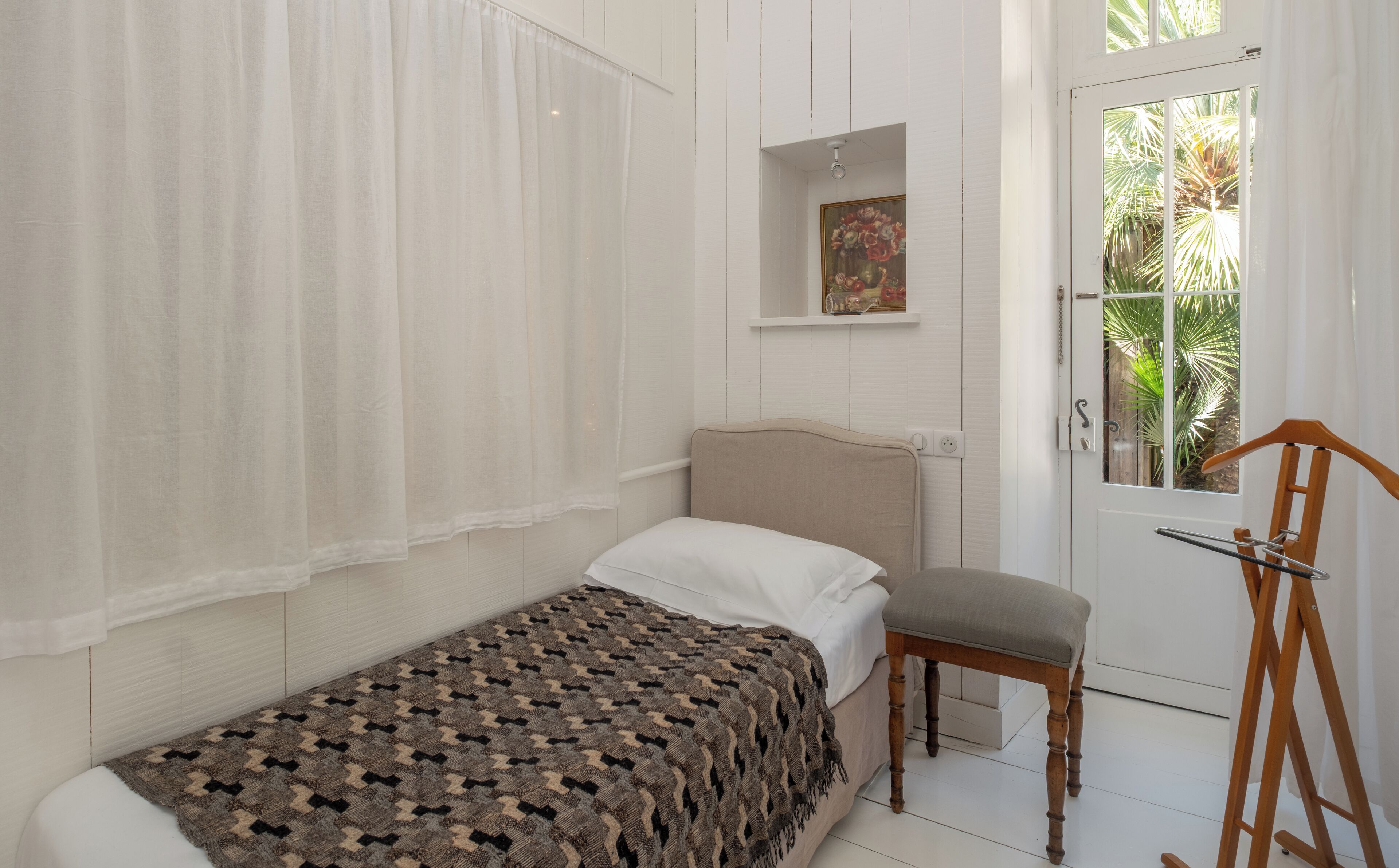 A small, neatly arranged bedroom with a single bed, a chair, a wooden ladder, and a door leading to a balcony with a view of greenery.