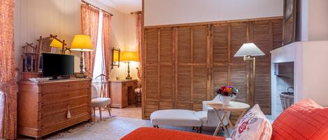 A cozy bedroom with a wooden dresser, a television, a desk with a lamp, a chair, and a bed with an orange blanket.