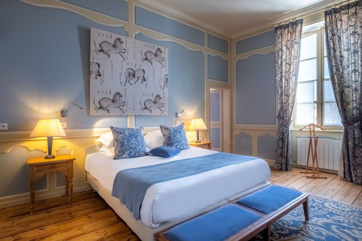 Une chambre avec un grand lit, deux tables de chevet avec des lampes, une œuvre d’art accrochée au mur et une fenêtre avec des rideaux.