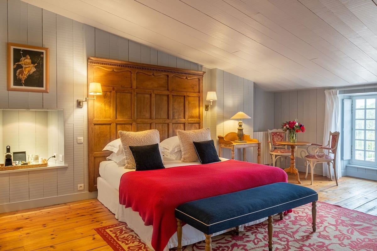 Une chambre à coucher avec une tête de lit en bois, une couvre-lit rouge, un banc, une petite table avec une lampe et une chaise sur laquelle est posé un vase de fleurs.