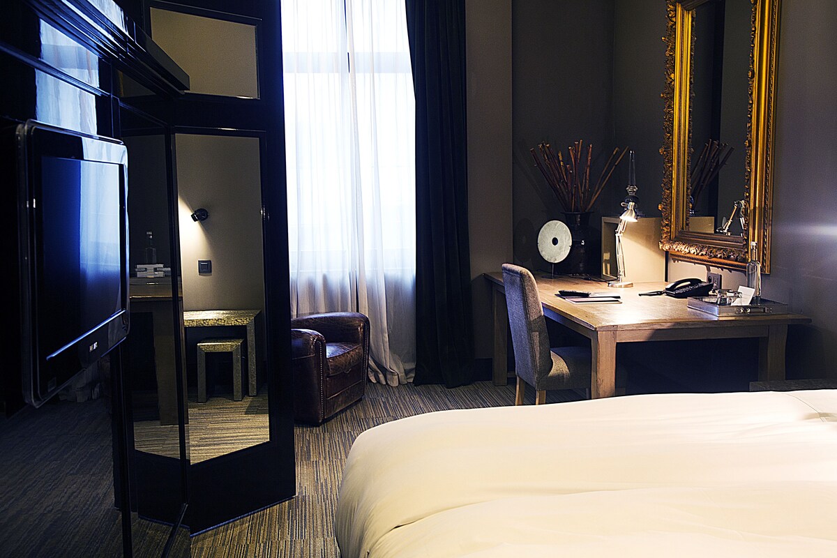 A hotel room with a bed, a desk with a chair, a mirror, and a window with curtains.