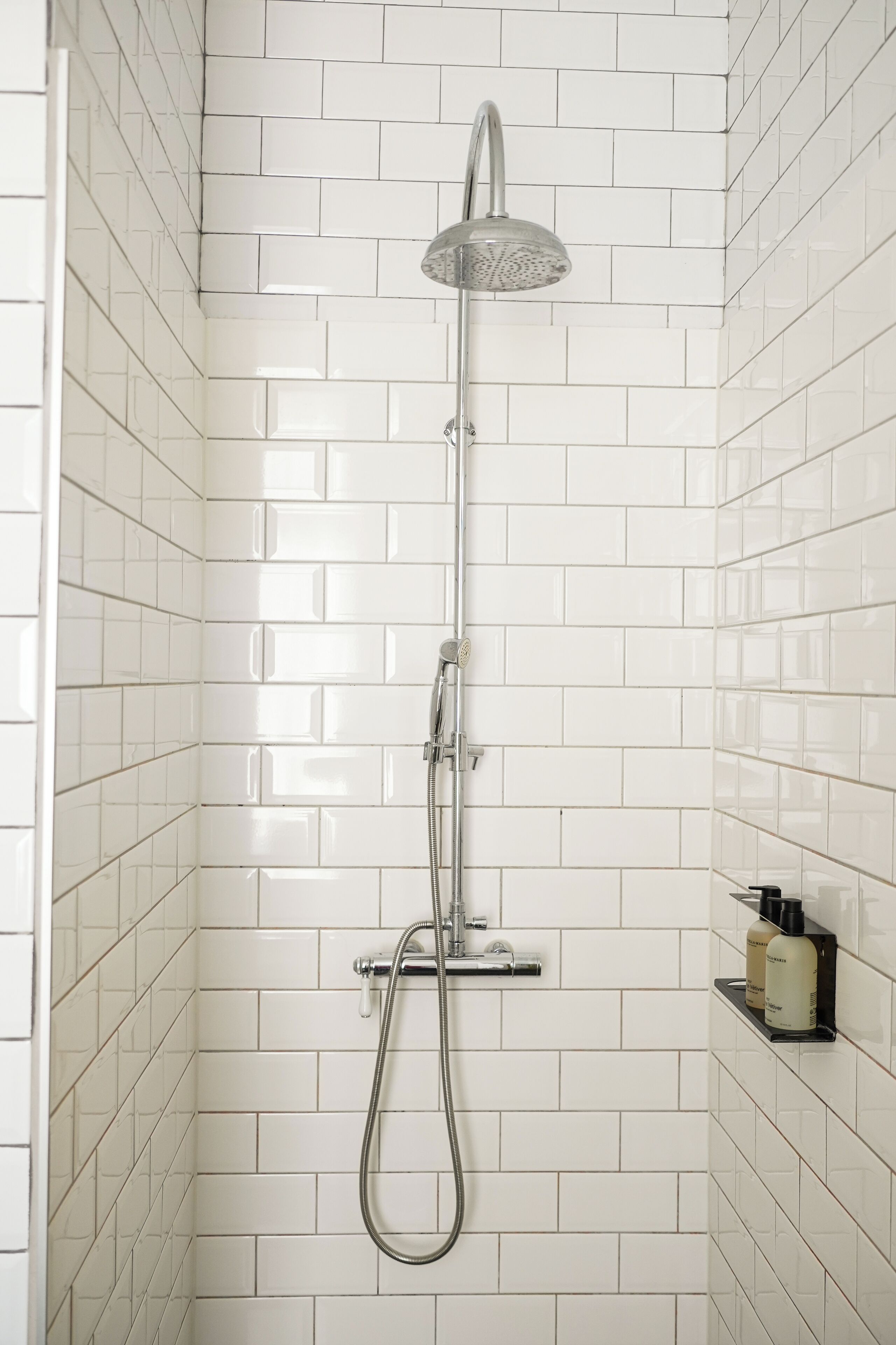 A shower with a handheld showerhead and a wall-mounted faucet, white tiled walls, and a small shelf with toiletries.