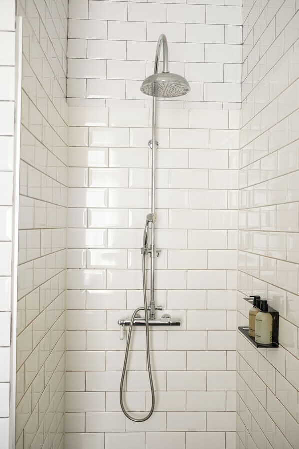 A shower with a handheld showerhead and a wall-mounted faucet, white tiled walls, and a small shelf with toiletries. - Les Nuits Hotel (Antwerp)