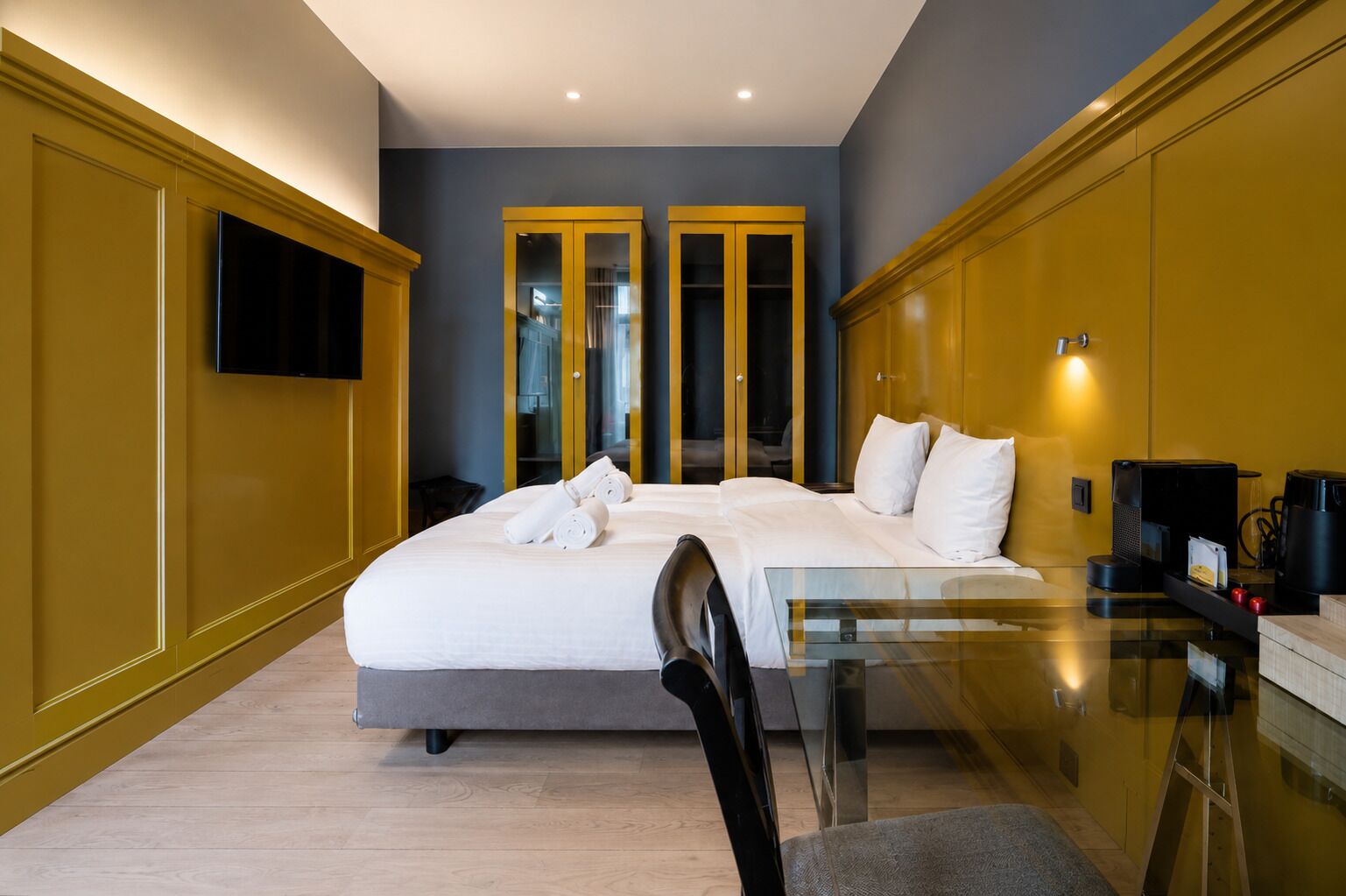 A hotel room with a bed, a glass table, a chair, a TV, and a coffee machine.