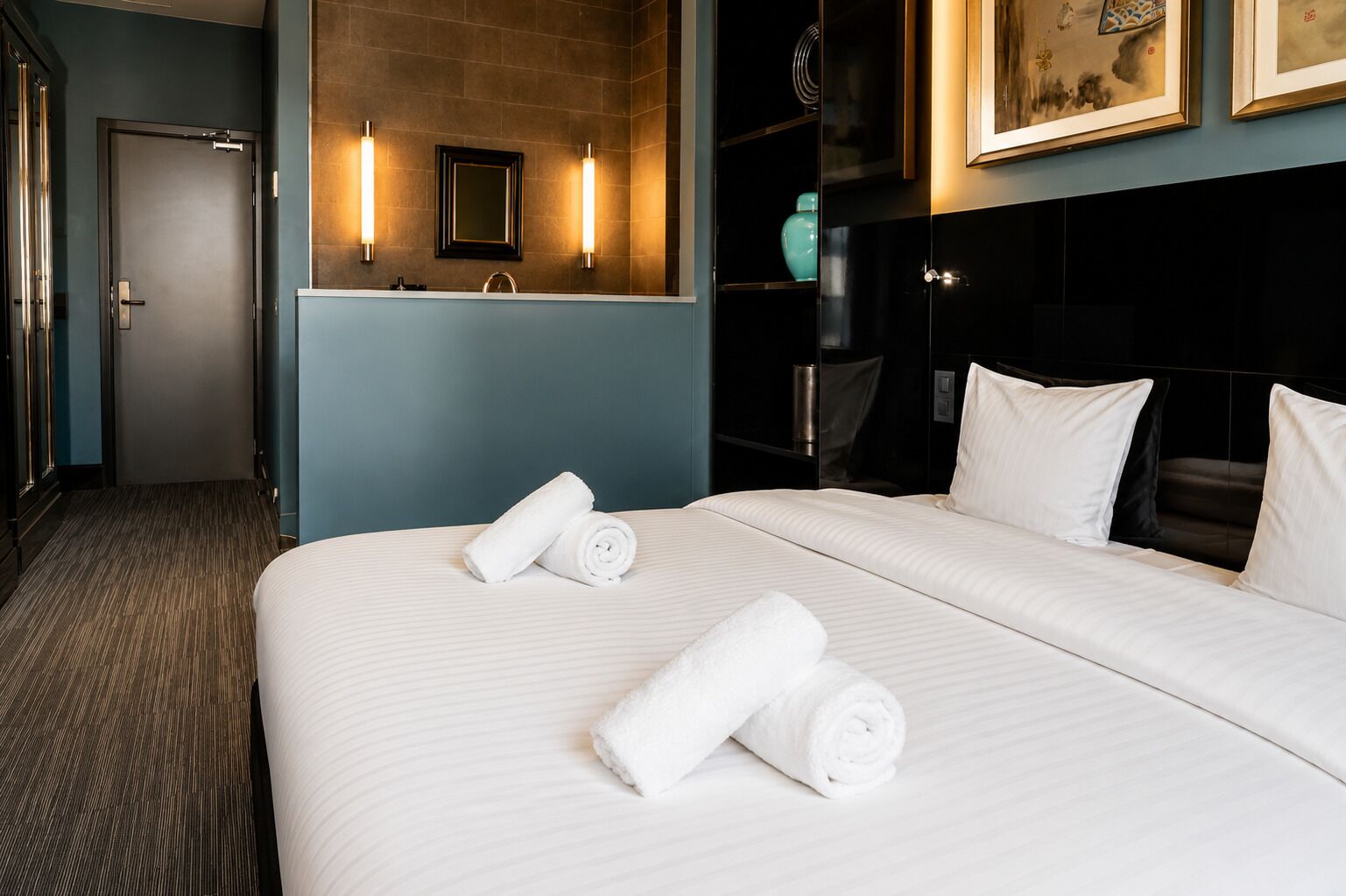 Ein Hotelzimmer mit einem großen Bett, zwei aufgerollten Handtüchern, einem Nachttisch, einem Spiegel und einem an der Wand befestigten Regal mit Dekorationen.