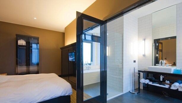 Habitación de hotel moderna con una cama grande, baño separado con puerta de cristal y vistas a la ciudad.