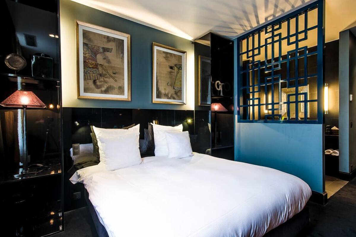 A modern hotel room with a large bed, two framed artworks on the wall, a decorative screen, and a nightstand with a lamp.