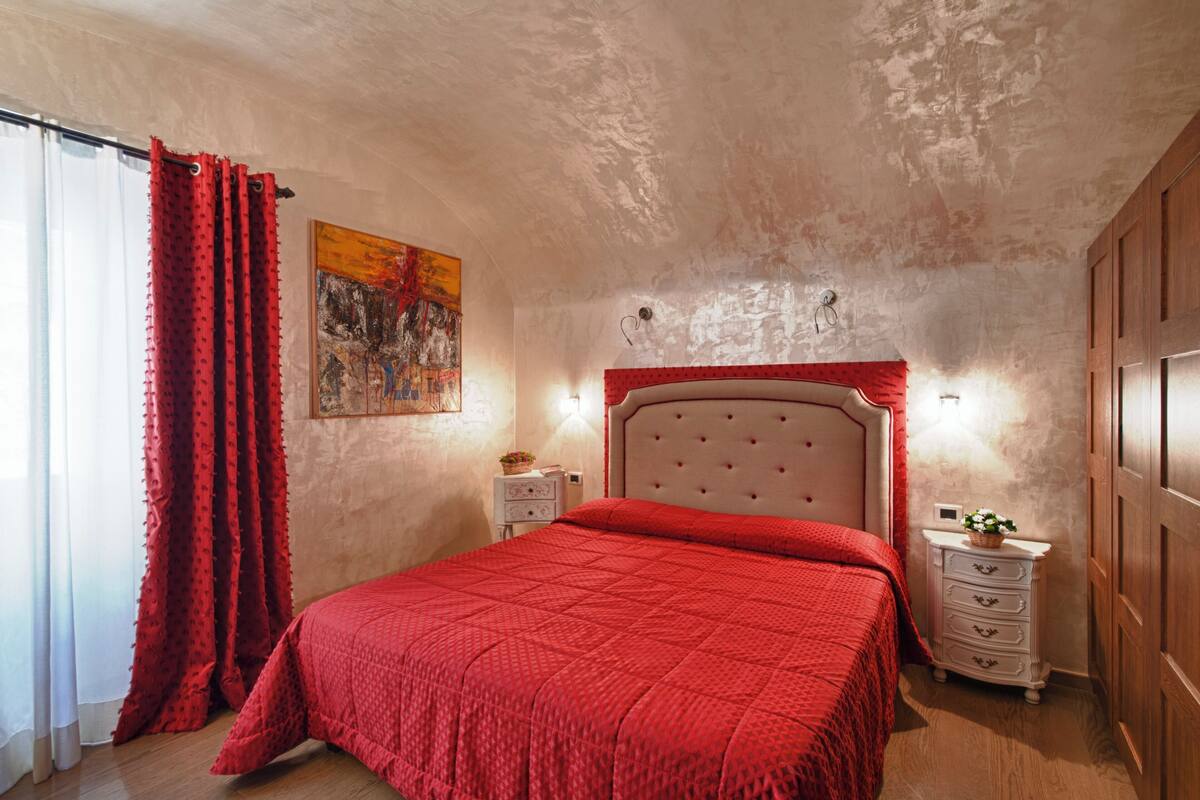 A bedroom with a red bedspread, a white nightstand with drawers, a red curtain, and a painting on the wall.