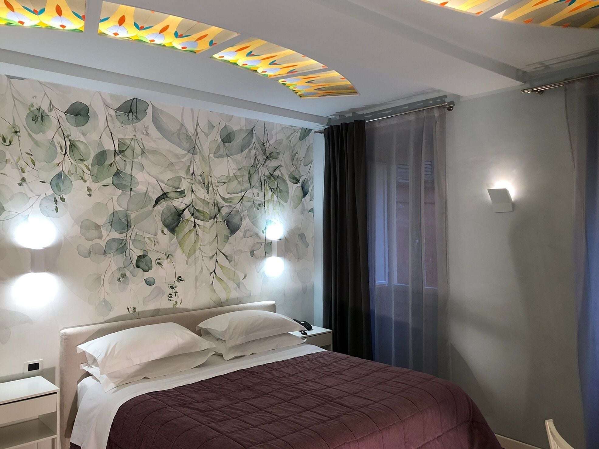 A hotel room with a bed, a nightstand, a wall with a leaf pattern, and a window with curtains.