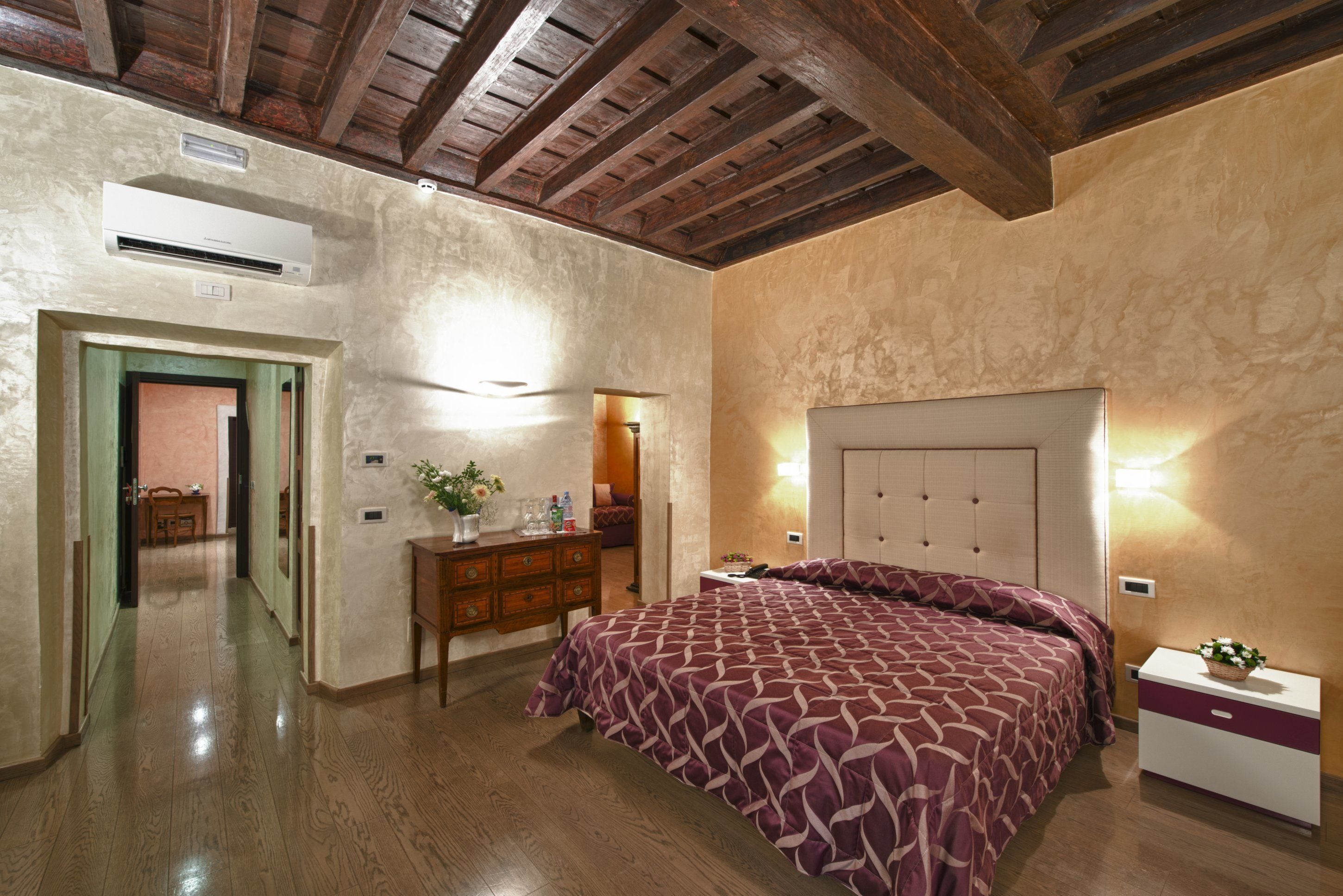 A bedroom with a wooden ceiling, a bed with a patterned bedspread, a dresser, and a nightstand with a plant.