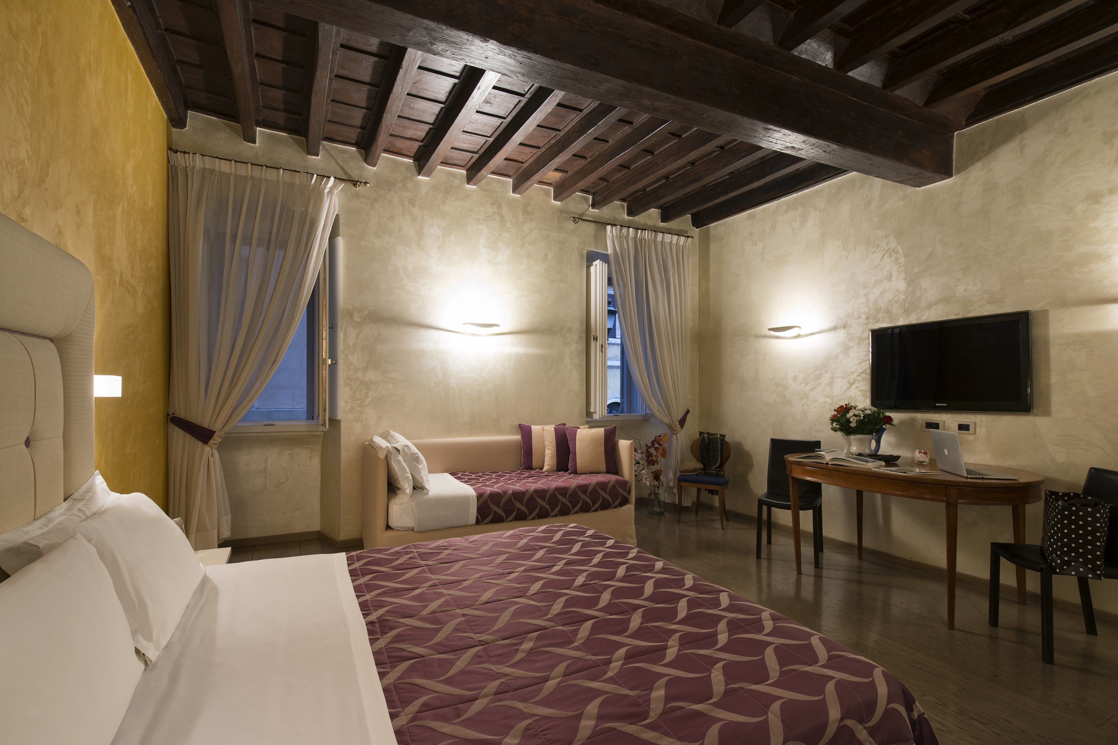 A hotel room with a large bed, a desk with a laptop, a TV, and a window with curtains.