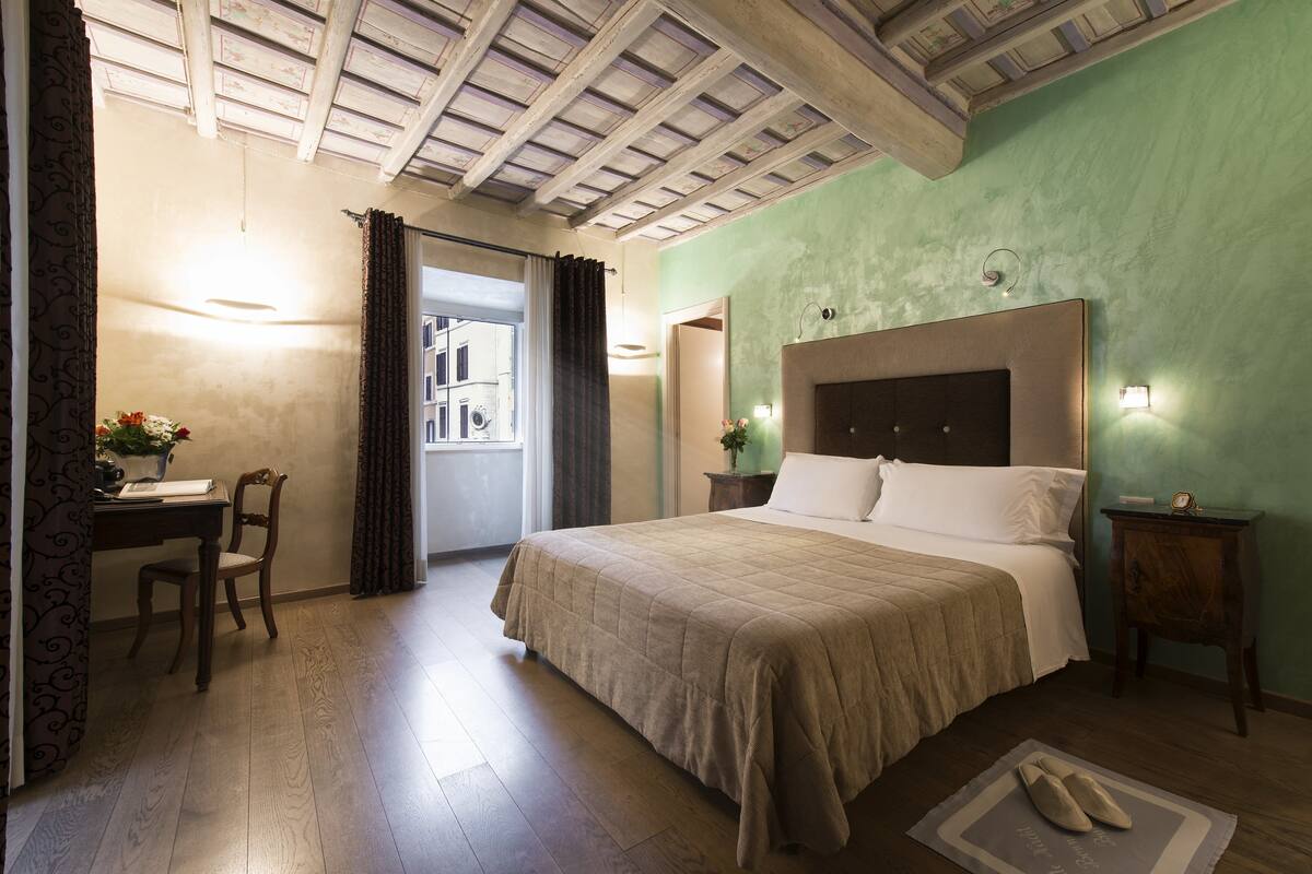 A bedroom with a large bed, a desk with a chair, a small table with a lamp, and a window with curtains.