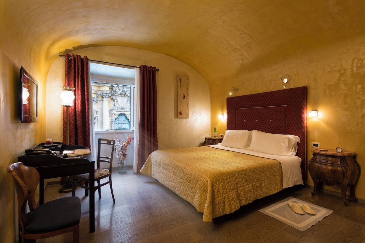 A hotel room with a bed, a desk with a chair, a piano, and a view of the outside.