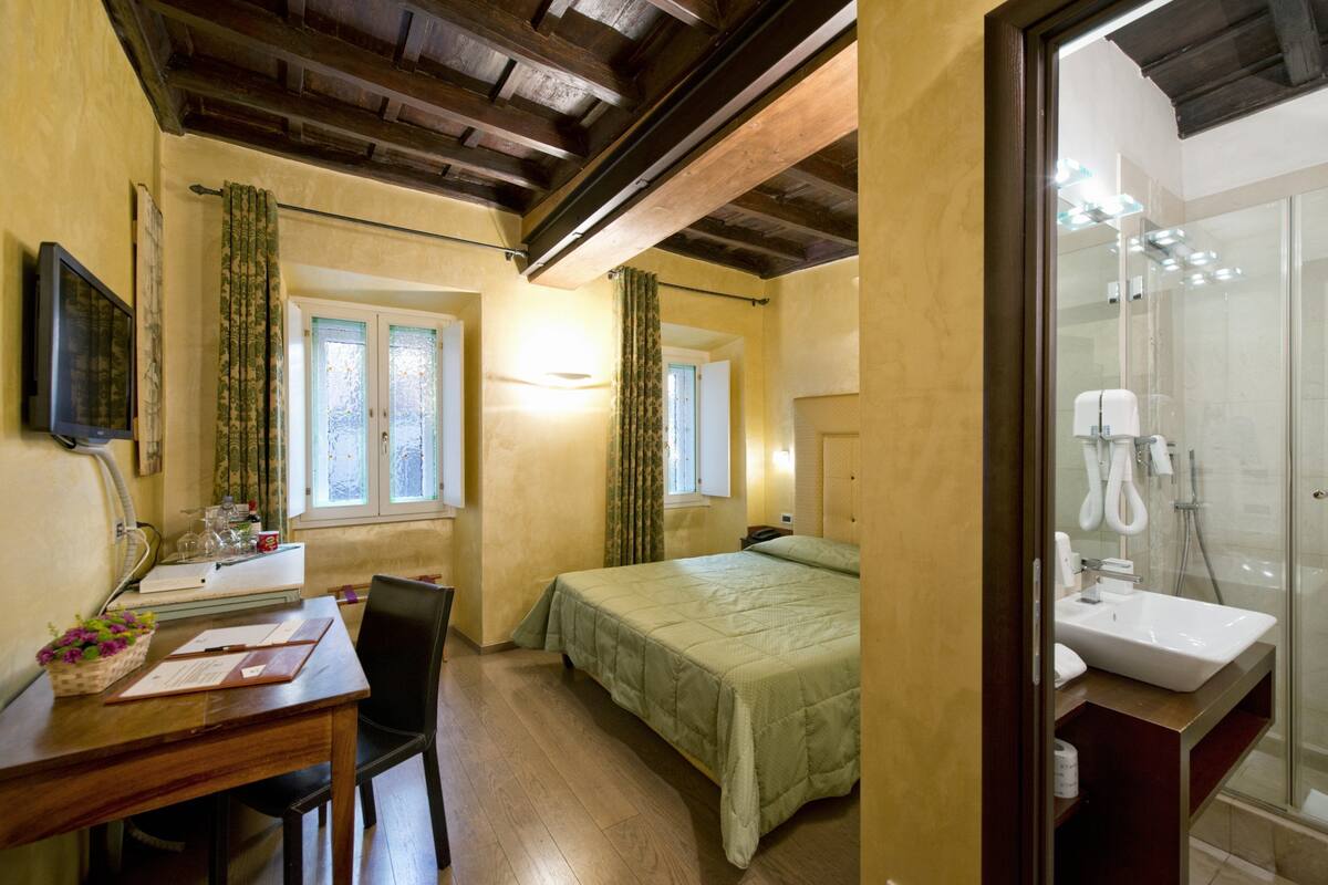 A bedroom with a bed, a desk with a chair, a window with curtains, and a bathroom with a sink and shower.