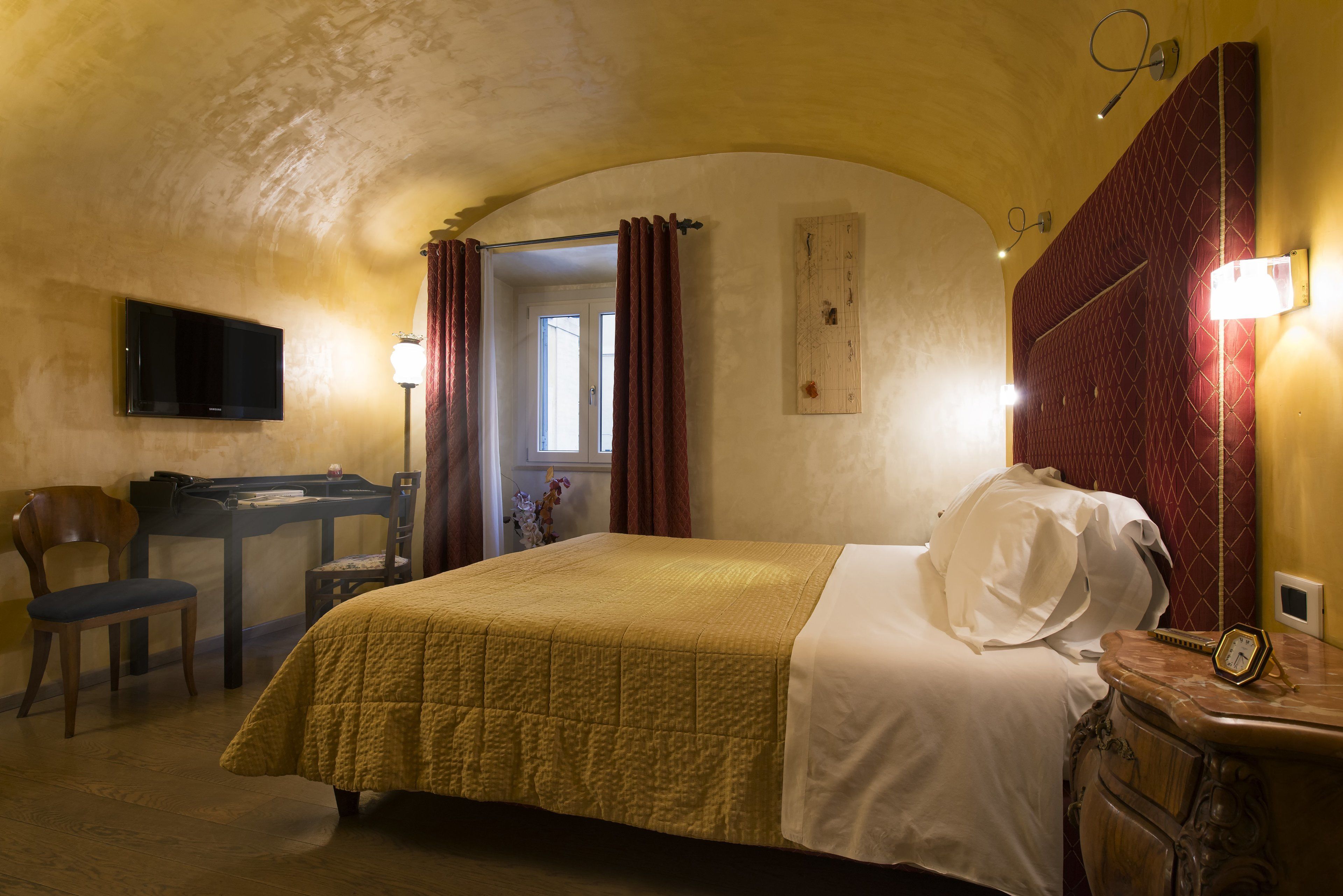 Ein Hotelzimmer mit einem Bett, einem Schreibtisch, einem Sessel, einem Fernseher und einer Uhr auf einem Nachttisch.