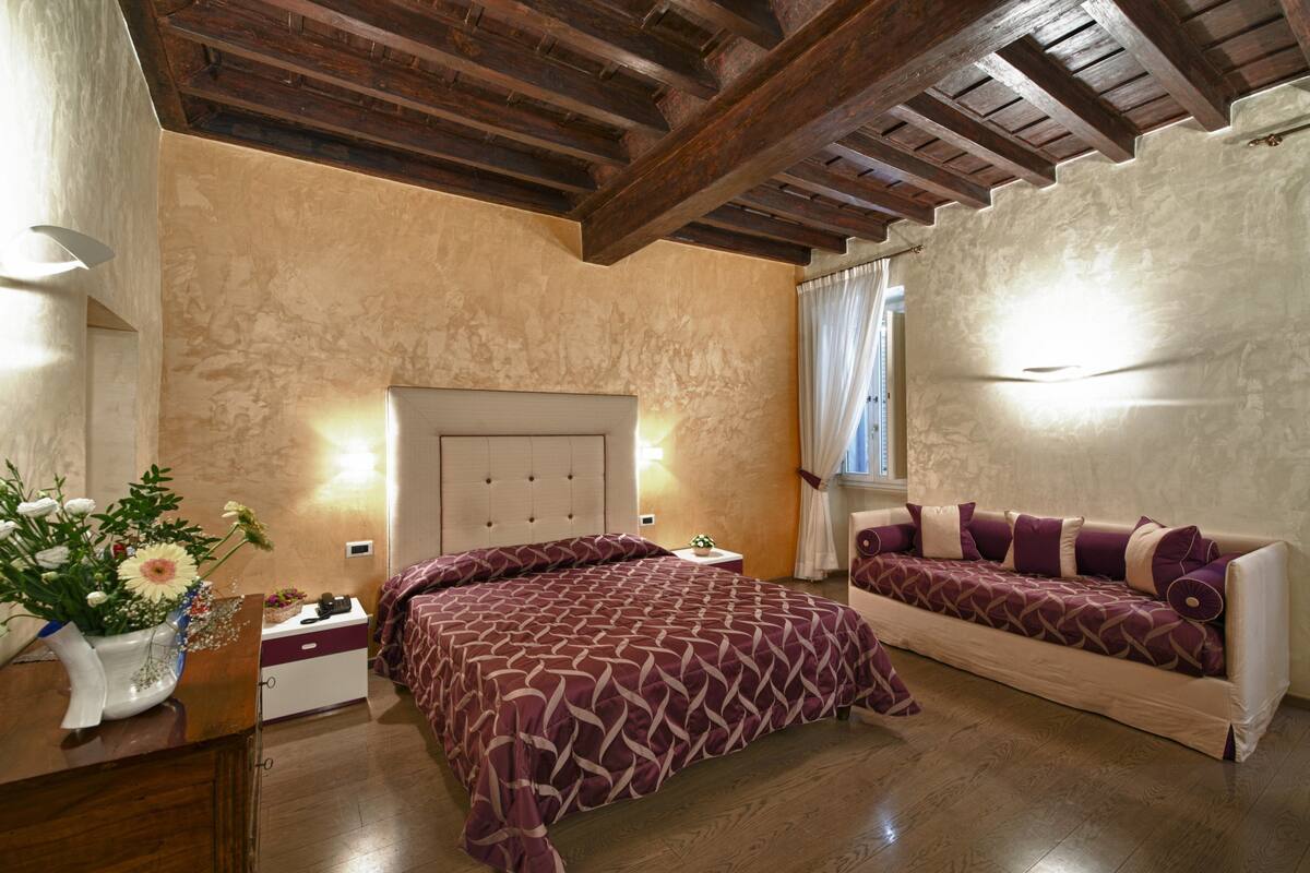 A bedroom with a bed, a sofa, a nightstand, and a vase with flowers.