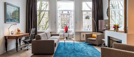 A modern living room with a sofa, armchair, and a blue rug. There is a desk with a chair, a lamp, and a framed picture on the wall.