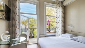 A hotel room with a bed, a glass table with a chair, a TV, and a balcony with a view of trees and a building. - Amstel Corner Hotel (Amsterdam)