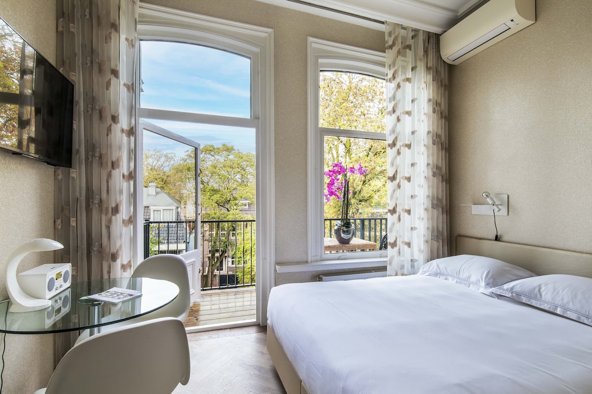 A hotel room with a bed, a glass table with a chair, a TV, and a balcony with a view of trees and a building.