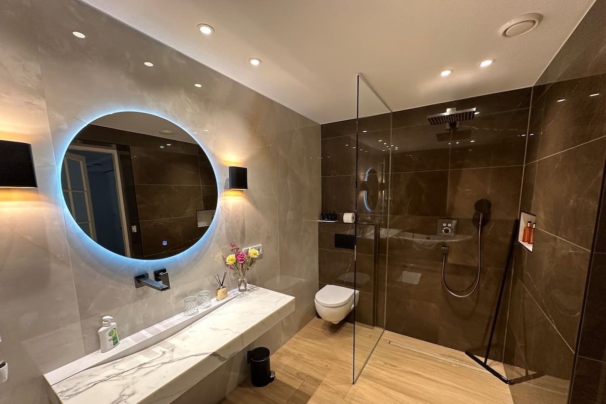A modern bathroom with a marble vanity, a round mirror with under-cabinet lighting, a glass-enclosed shower, and a toilet.