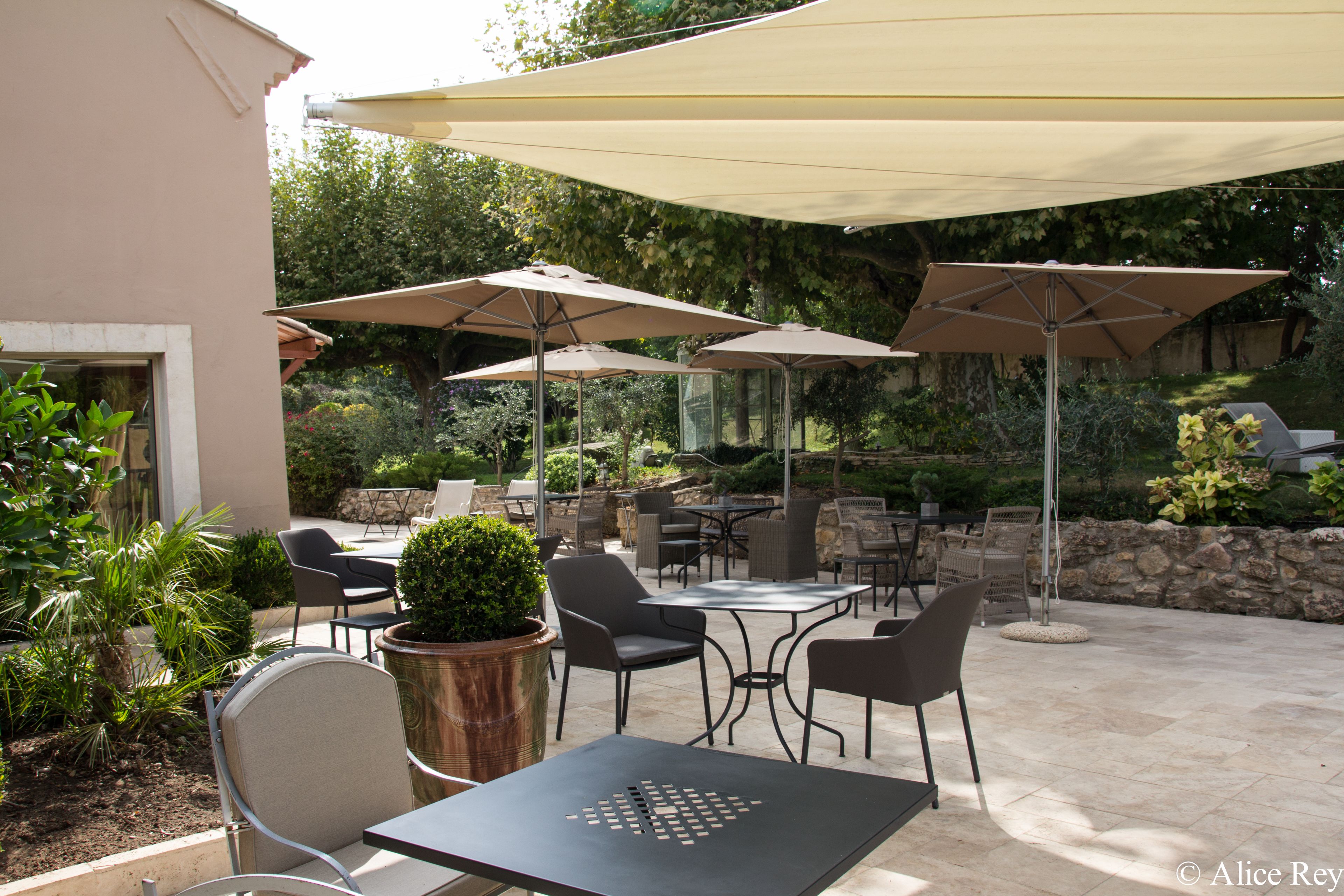 An outdoor patio area with multiple tables and chairs, shaded by large umbrellas, surrounded by greenery and stone walls.