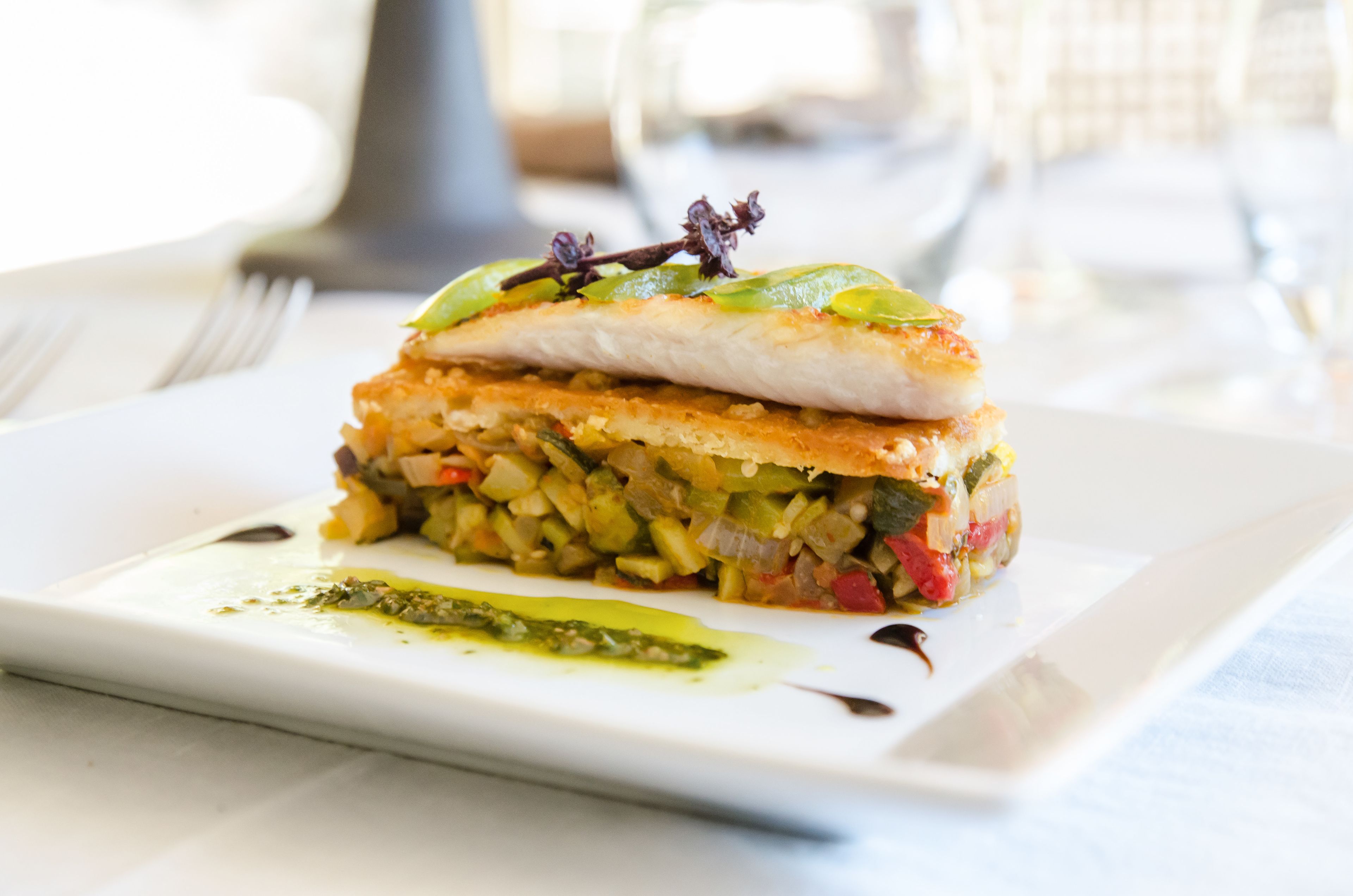 Um prato com camadas, possivelmente um sanduíche ou um gratinado, sobre um prato branco, acompanhado de um molho verde e um fio escuro.