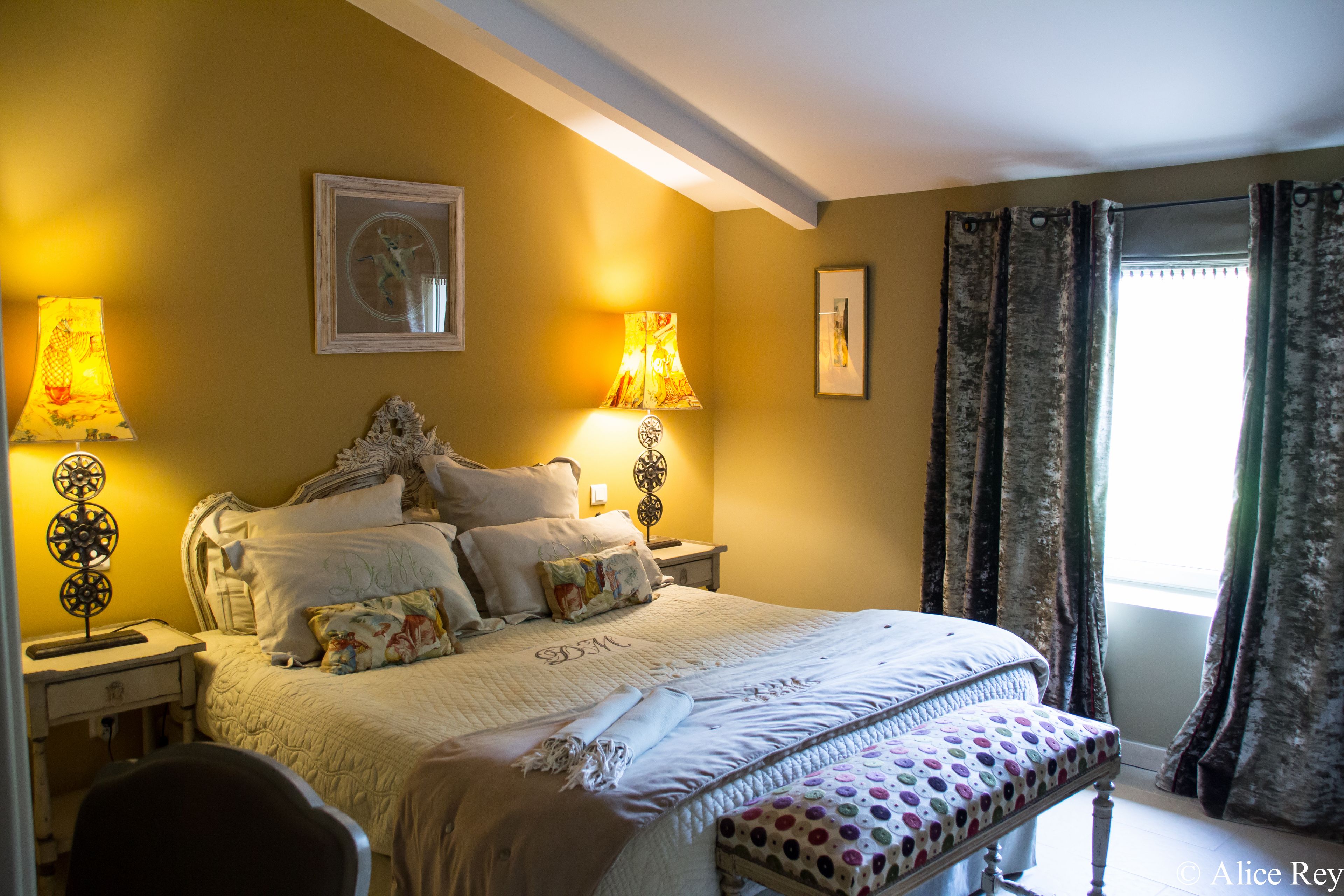 Une chambre bien éclairée, avec un grand lit, deux lampes de chevet, une table de nuit et une fenêtre avec des rideaux.