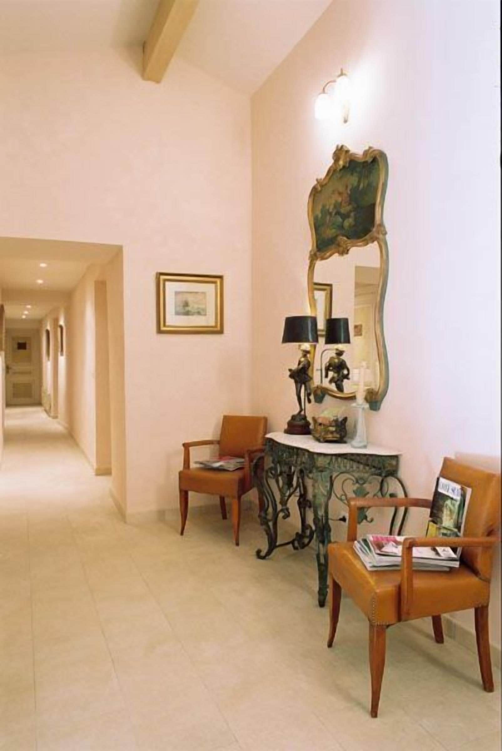 Um corredor com duas cadeiras de madeira, uma mesa consola com um abajur e um espelho grande na parede.