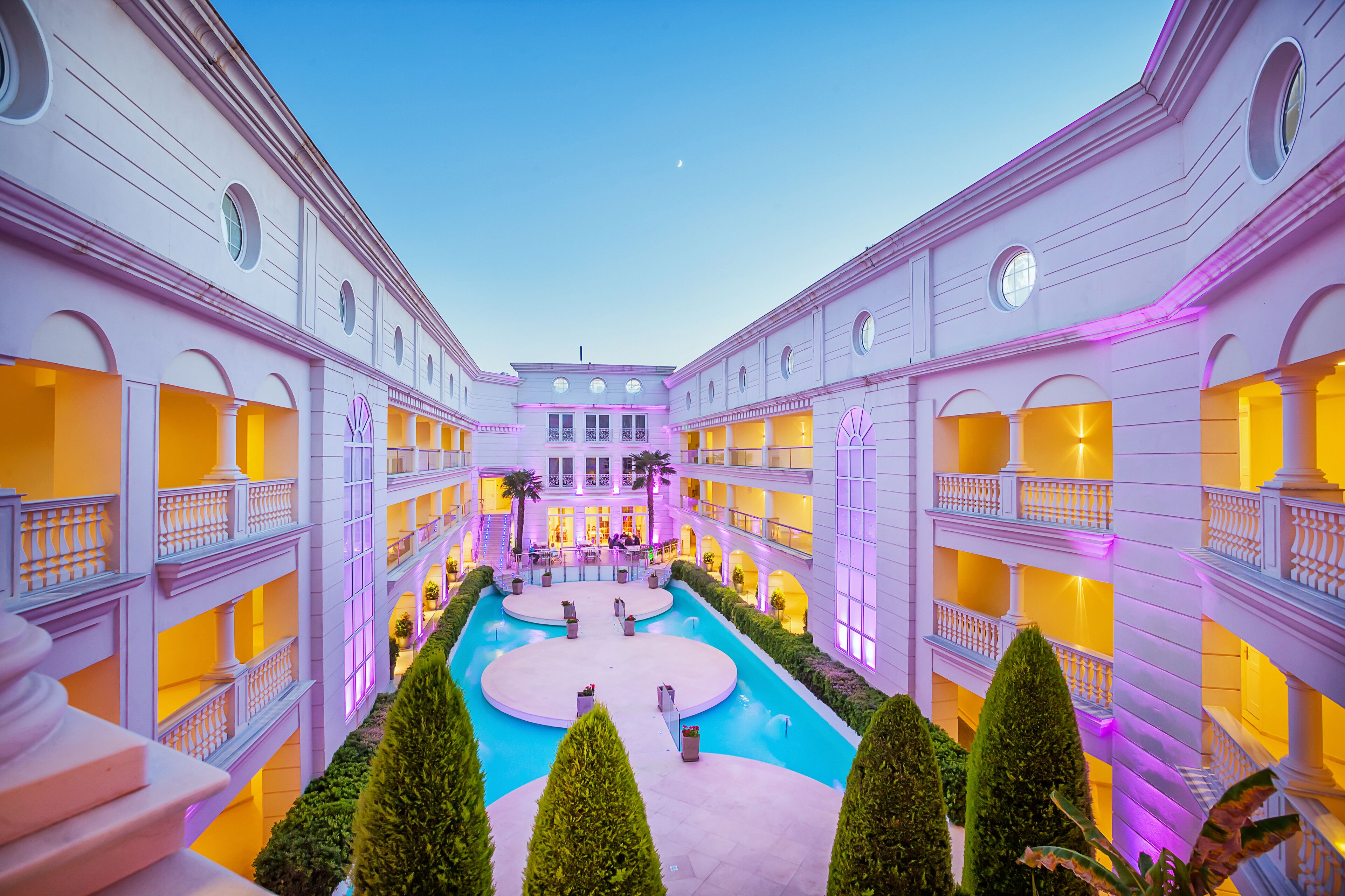A multi-level hotel atrium with a central pool, balconies, and a clear sky.