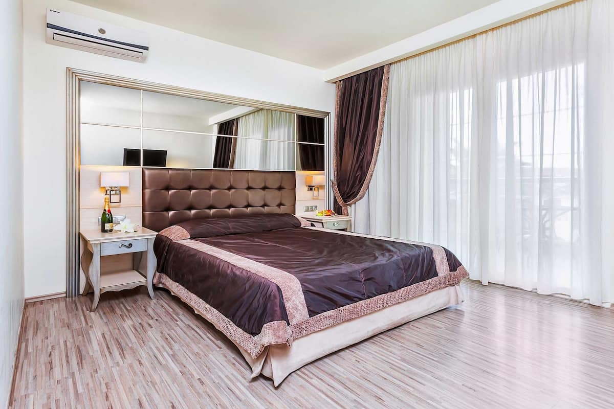 Un dormitorio amplio con una cama grande, una mesita de noche con lámpara, un espejo y una ventana con cortinas.
