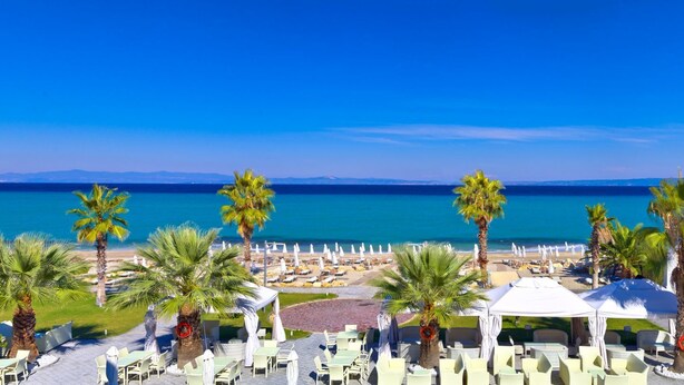 A beachside resort with outdoor seating and palm trees, overlooking a clear blue sea.