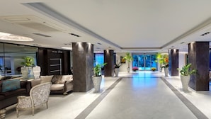 A modern hotel lobby with a central pathway, seating areas on both sides, and large windows with a view of greenery.