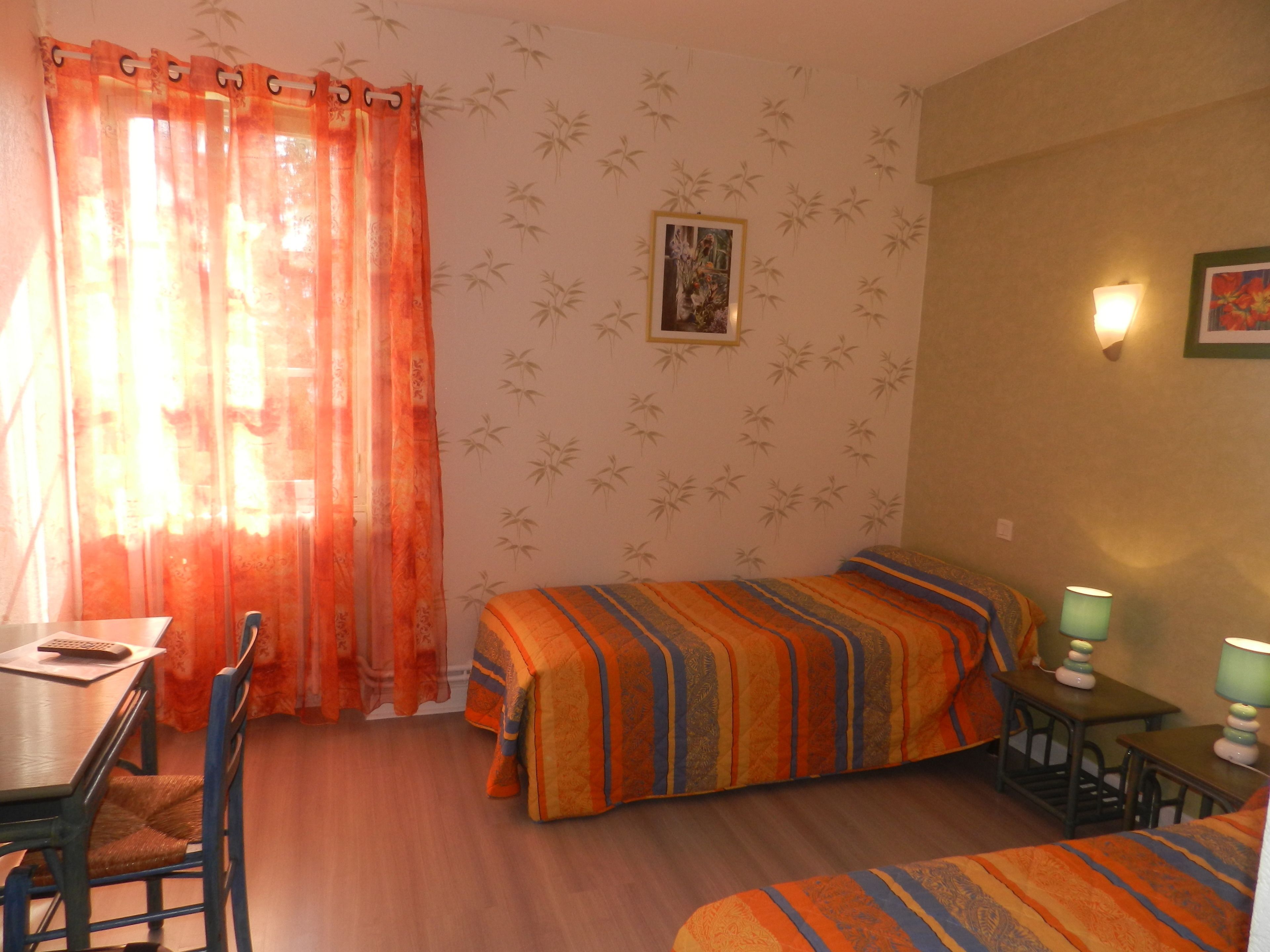 A room with two beds, a desk, and a chair. There is a window with orange curtains, a wall with bamboo wallpaper, and a framed picture on the wall.