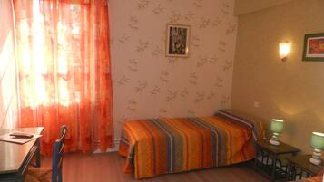 A room with two beds, a desk, and a chair. There is a window with orange curtains, a wall with bamboo wallpaper, and a framed picture on the wall.