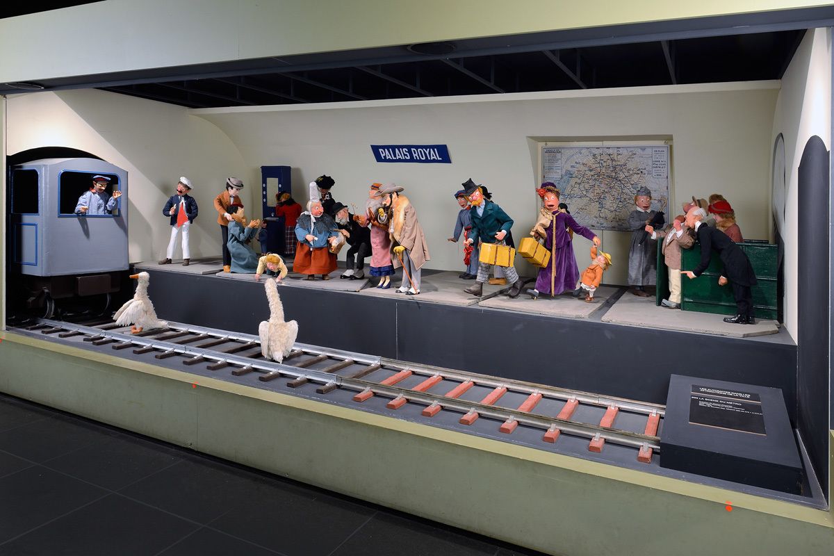 A model train exhibit with figures dressed in historical attire, a sign reading "PALAIIS ROYAL," and a map on the wall.