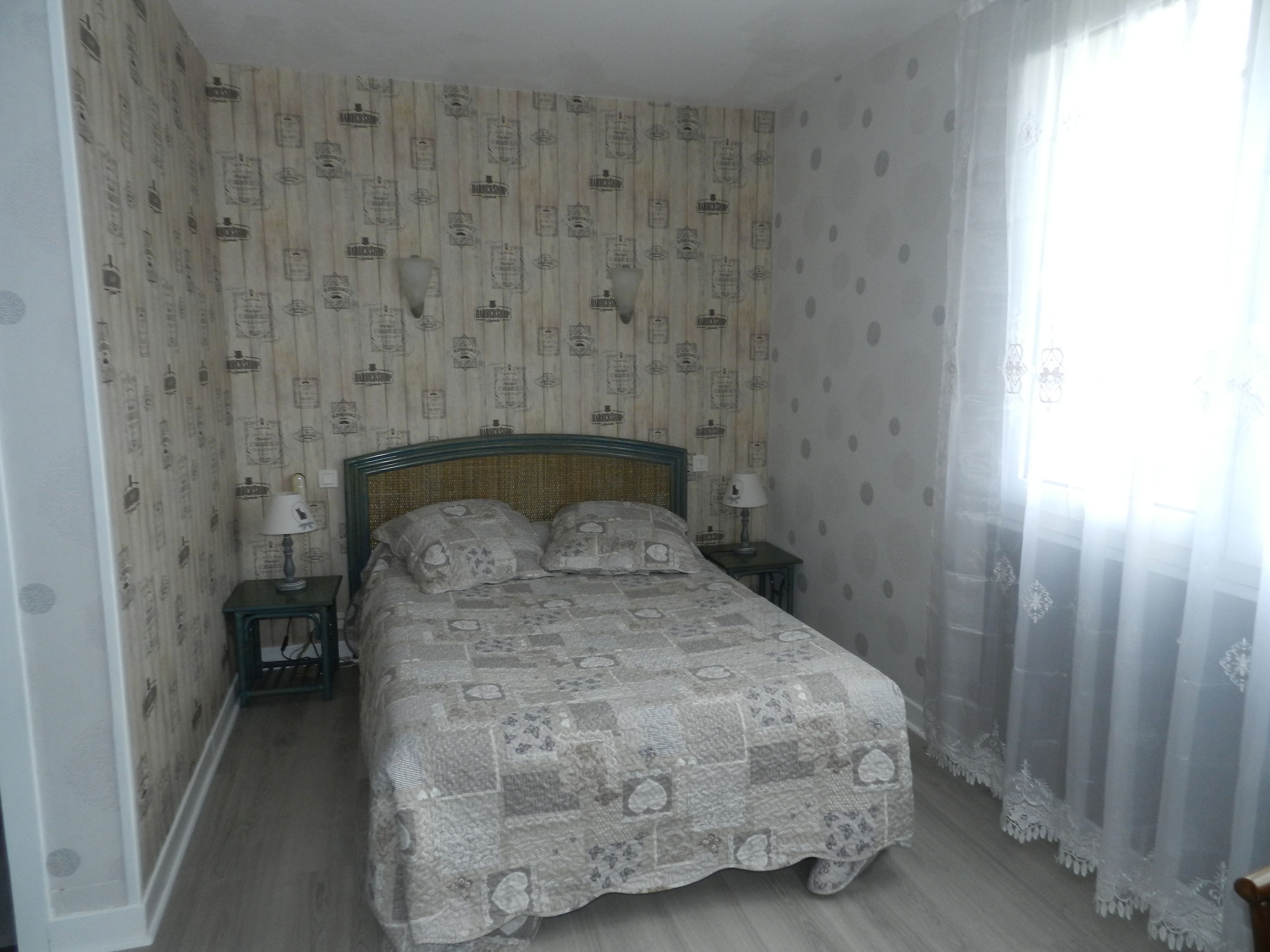 A bedroom with a bed, two bedside tables, a window with sheer curtains, and patterned wallpaper.
